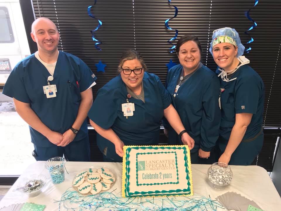4 people standing next to table holding cake